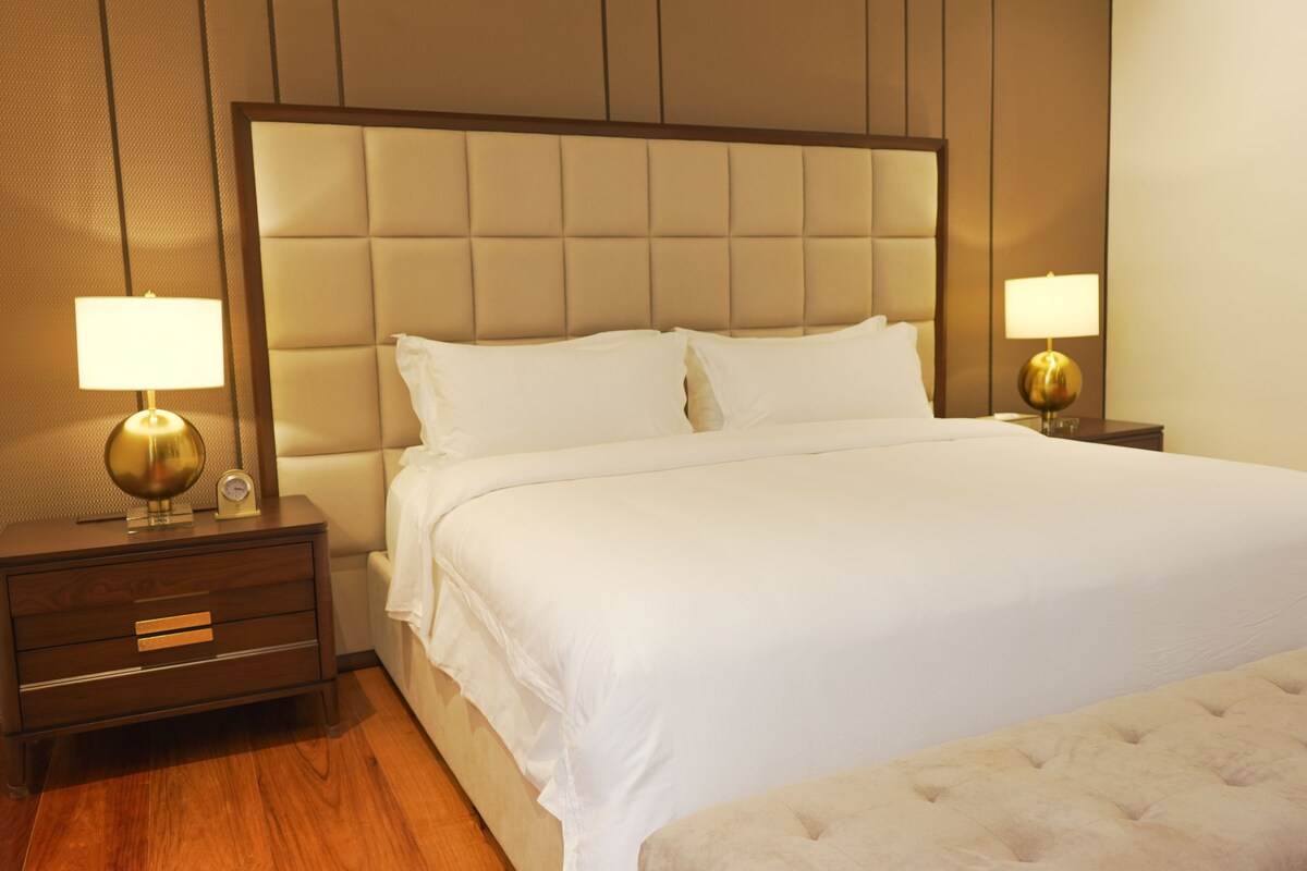 Una habitación de hotel con una cama grande, dos mesitas de noche con lámparas, una mesita auxiliar de madera y suelo de madera.