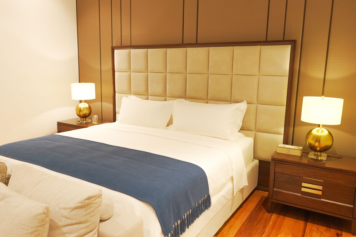 Habitación de hotel con una cama grande, dos mesitas de noche con lámparas, piso de madera y cabecera acolchada.