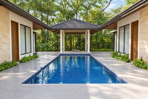 Una piscina moderna con solárium empedrado, rodeada de exuberante vegetación y un área de descanso cubierta.