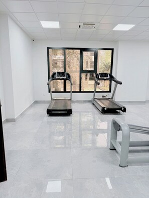 A fitness room with two treadmills and a bench, large windows with a view of trees, and a tiled floor. - Rooftop Pool with Riverview 3 bedroom apartment (Kinshasa)