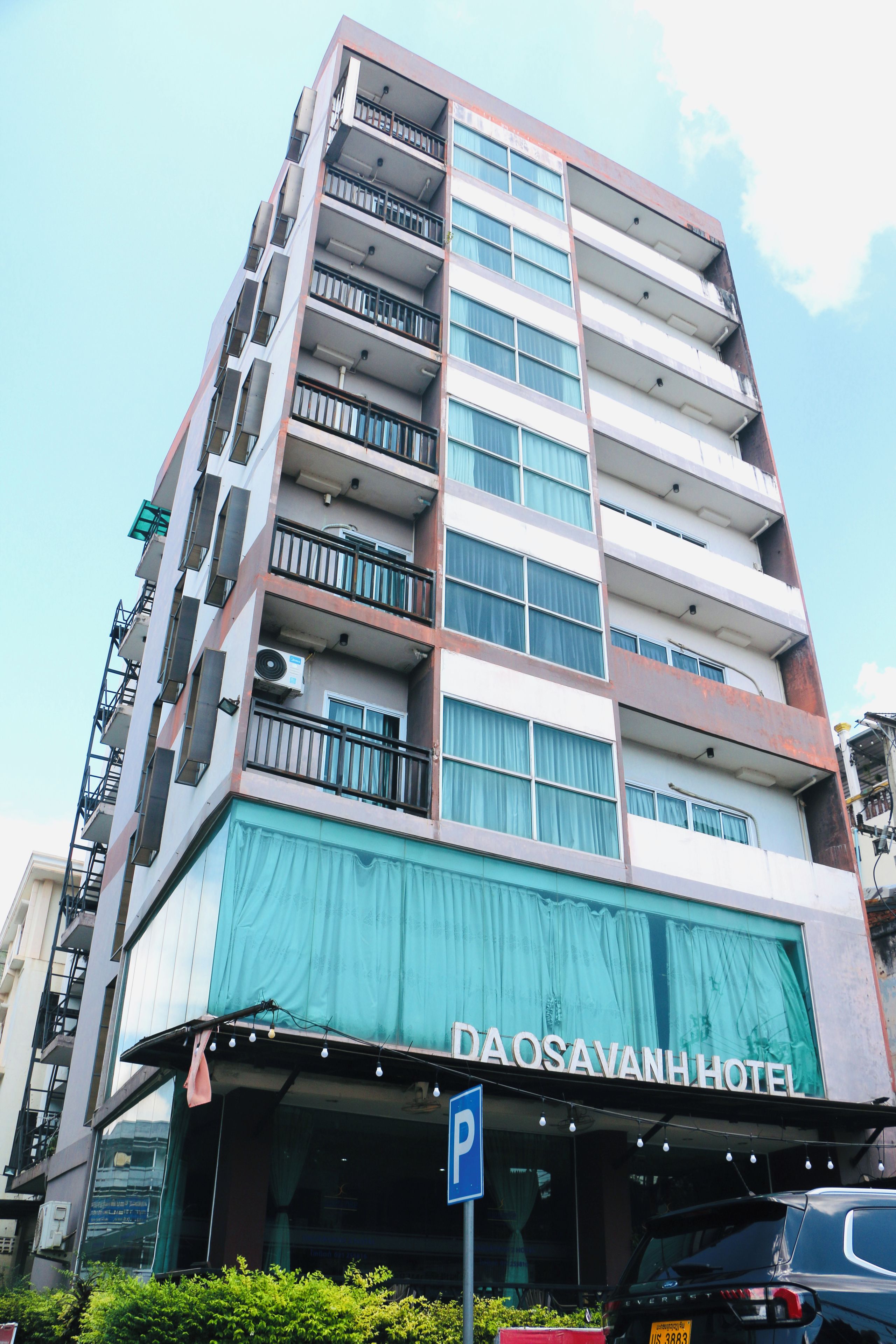 Un hotel de varios pisos con balcones en cada planta, un cartel visible que dice 'DAOSAVANH HOTEL' y un letrero de estacionamiento que indica una zona de aparcamiento.