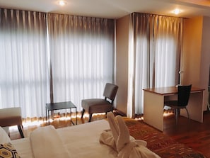 Una habitación de hotel con una cama, dos sillas, un escritorio y un ventanal con cortinas.