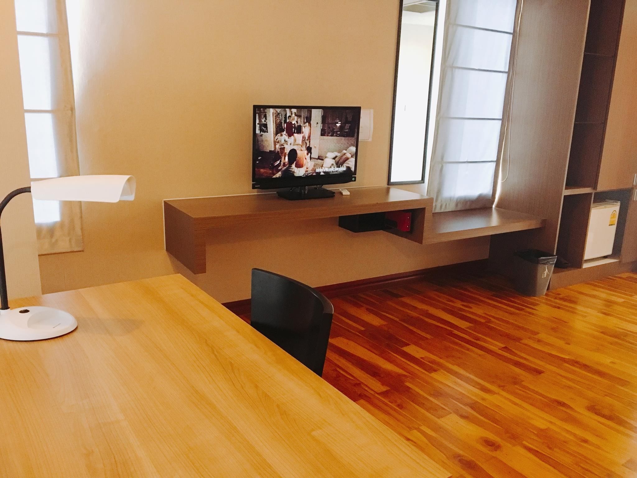 Una habitación con piso de madera, un televisor sobre un mueble, una lámpara de escritorio blanca y una silla.