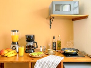Eine Küche mit Mikrowelle, Mixer, Kaffeemaschine und einer Bratpfanne auf dem Herd. Es sind Früchte, ein Glas Orangensaft und ein Teller mit Avocado-Scheiben vorhanden.
