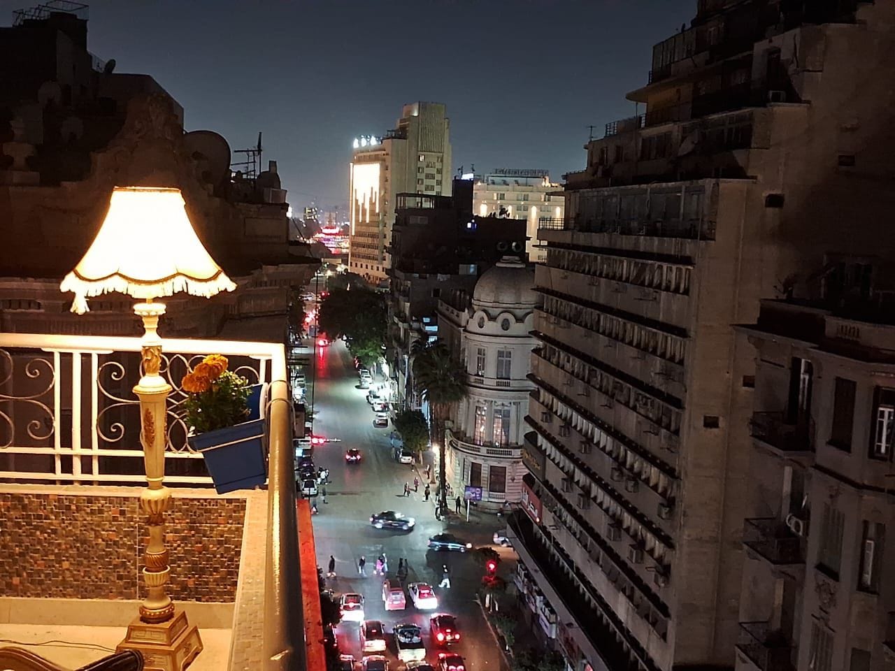 A nighttime cityscape with buildings, a busy street, and a balcony with a lamp.