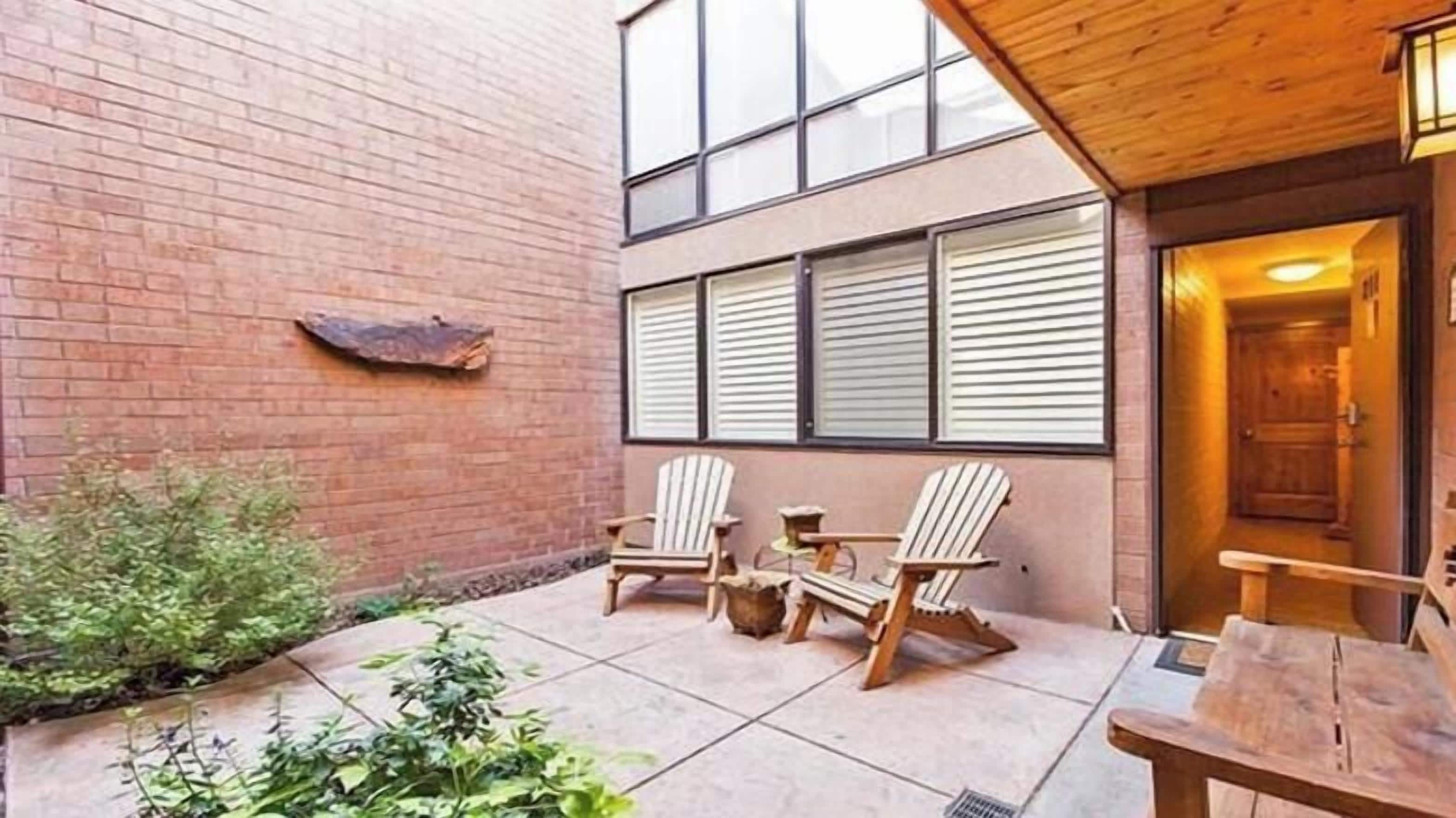 Une terrasse avec deux fauteuils Adirondack, une petite table et un mur en briques percé d’une fenêtre.