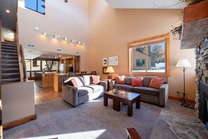 A spacious living area with a fireplace, a kitchen in the background, and a staircase leading to an upper level. - Park Ave 224 | Heated Pool | Private Hot Tub | Near PCMR | Laundry | Walk to Dining | Free Bus (Park City)
