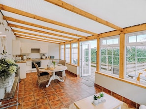Eine Küche mit Holzbalken, einem Esstisch und Blick auf einen Garten durch große Fenster.
