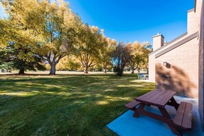Una mesa de picnic en un área con césped, árboles y un edificio de ladrillo al fondo.