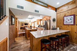 A kitchen with wooden cabinets, a stainless steel refrigerator, a bar area with stools, and a sign that reads 'WELCOME TO THE MOUNTAINS'. - Park Ave 236 | Heated Pool | Hot Tub | Mtn Views | Trail-Side by Golf Course | Laundry | Near PCMR (Park City)