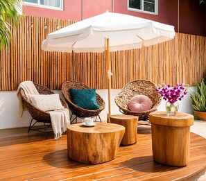 Ein Sitzbereich im Freien mit Korbstühlen, runden Holztischen und einem weißen Sonnenschirm.