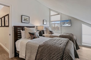 Quarto com duas camas, janela com persianas e quadros nas paredes.