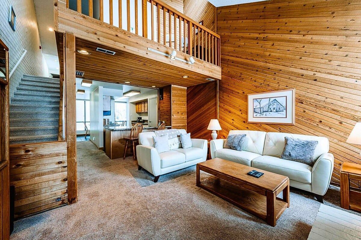 A spacious living area with a staircase, wooden walls, a sofa, a coffee table, and a dining area in the background.