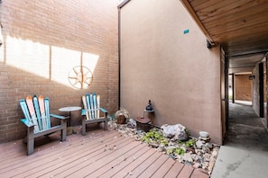 A wooden deck with Adirondack chairs and a small round table, a brick wall with a decorative wheel, and a stone pathway leading to a building. - Park Ave 297 | Heated Pool | Hot Tub | No Stairs | Private Patio & Grill | Fireplace | Free Bus (Park City)