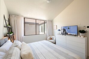 Ein Schlafzimmer mit großem Fenster, einem Bett mit gestreifter Bettwäsche, einer weißen Kommode und einem Flachbildfernseher sowie einem Sessel.
