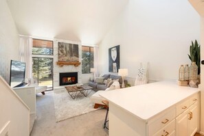 A modern living room with a fireplace, a sofa, a coffee table, and a kitchen area with cabinets and a countertop. - Park Ave 299 | Heated Pool | Hot Tub | Remodeled End Unit | Fire Table Atrium | Patio & Grill (Park City)