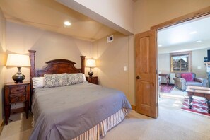 A bedroom with a large bed, two bedside tables with lamps, a wooden headboard, and a door leading to a living area with a sofa and a window. - Jupiter Inn 10 | Community Hot Tub | Gas Fireplace | A/C | Laundry | Walk to Dining | Free Bus (Park City)