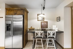 A kitchen with a stainless steel refrigerator, wooden cabinets, a bar stool, and a pendant light. - Jupiter Inn 18 | Mtn View Balcony | 2 Living Areas | Fireplace | Hot Tub | A/C | Free Bus (Park City)