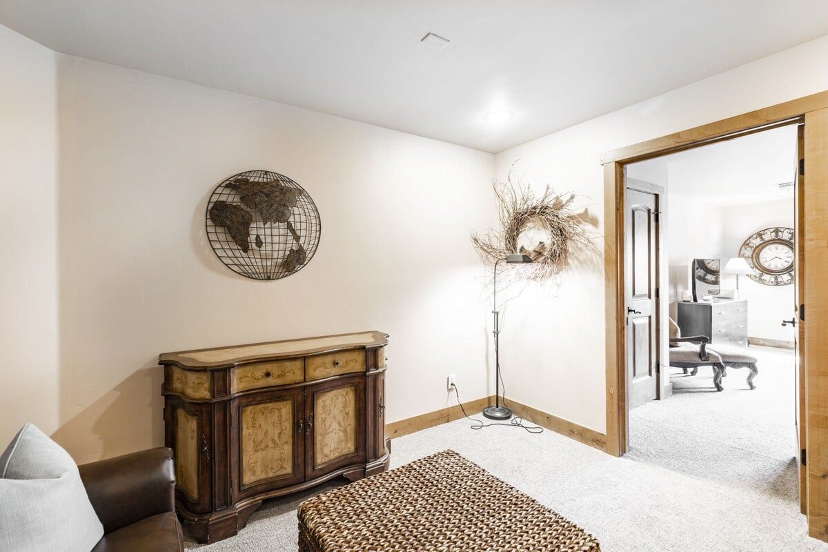 A room with a wooden cabinet, a woven ottoman, a globe artwork, a tall floor lamp, and a doorway leading to another room.