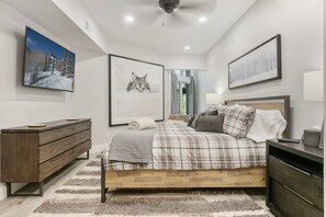 Quarto com cama, cômoda, mesa de cabeceira, abajur e quadros nas paredes.