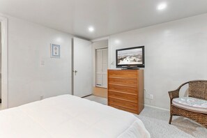 Une chambre à coucher comprenant un lit, une commode, un téléviseur posé sur un meuble, un fauteuil en osier et une porte donnant sur une autre pièce.