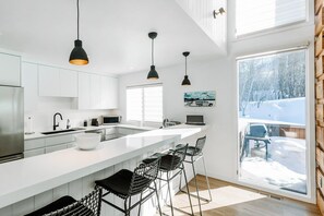 A modern kitchen with white cabinetry, a large island with bar stools, pendant lighting, and a view of a snowy landscape through a large window. - Three Kings 80 | Walk to Three Kings Lift | Heated Pool | Hot Tub | Remodeled | Original Artwork (Park City)