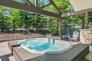 A hot tub with a wooden deck, surrounded by trees and a stone wall. - Jupiter Inn 11 | Community Hot Tub | Stone Fireplace | A/C | Laundry | Near Golf Course | Free Bus (Park City)