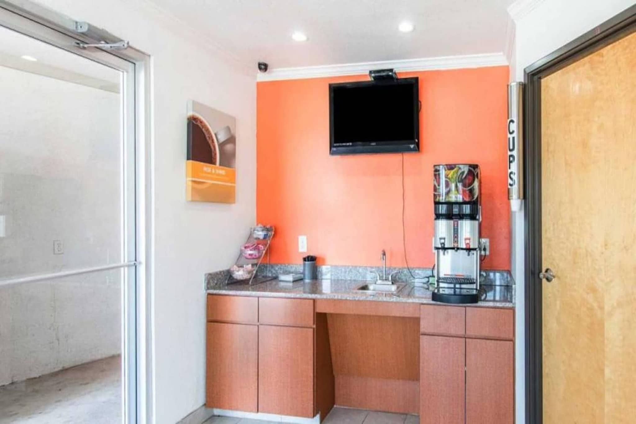 A small kitchenette with a coffee machine, a television mounted on the wall, and a door labeled 'Cups'.