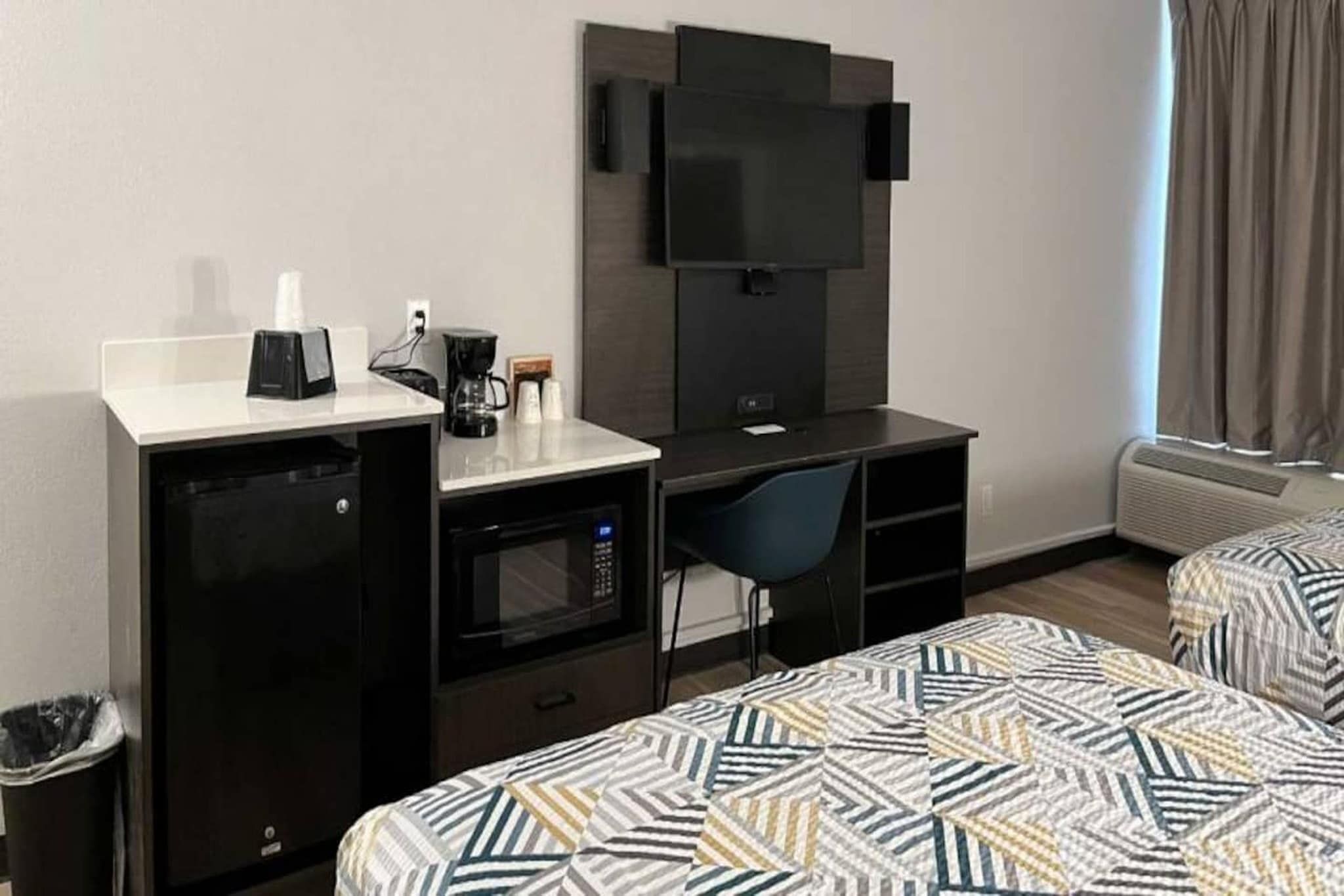 A hotel room with a kitchenette, a flat-screen TV, a desk, a chair, and two beds with patterned bedding.