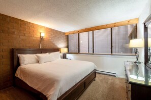A bedroom with a brick wall, a wooden headboard, a large bed, a nightstand with a lamp, a dresser, and a window with blinds. - Cozy Park City 1BR, Short Walk to the Resort (Park City)