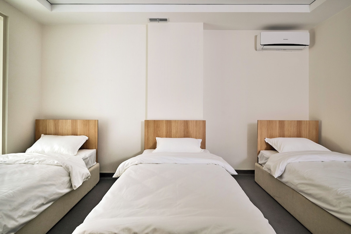 A hotel room with three single beds, a ceiling vent, and a plain wall.