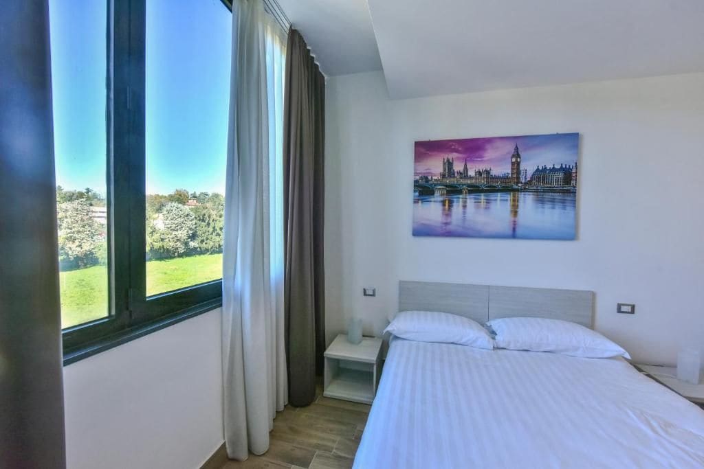 Habitación de hotel con un ventanal grande, una cama, una mesita de noche y un cuadro de un paisaje urbano.