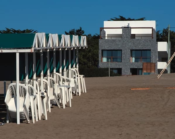 Un edificio de diseño moderno, un área de descanso techada con sillas y suelo arenoso.