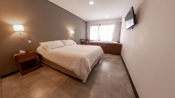 Habitación de hotel con cama, lámparas de noche, televisor montado en la pared y una ventana con cortinas.
