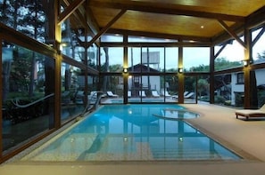 Una piscina interior con paredes de vidrio transparente, vigas de madera y sillas de descanso.