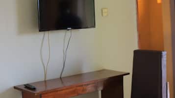 A room with a flat-screen TV mounted on the wall, a wooden desk with a chair, and a wall-mounted light fixture.