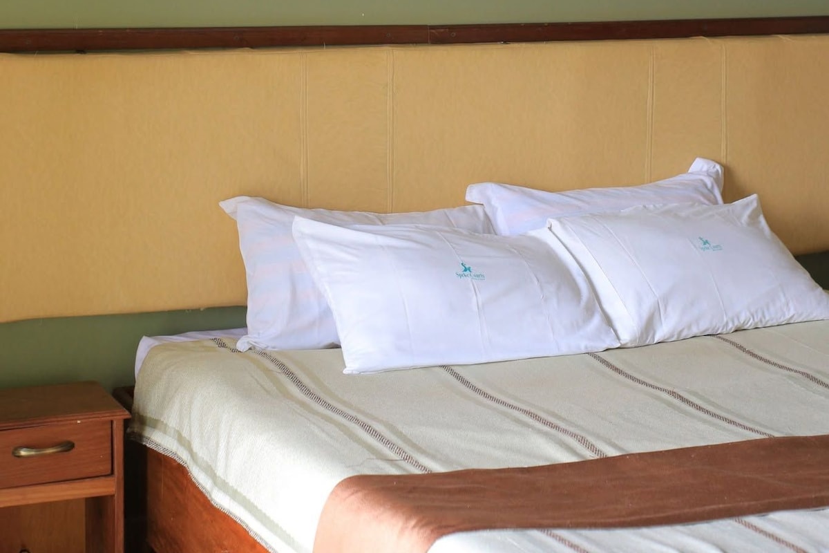 A hotel room with a bed, pillows, a nightstand, a framed picture on the wall, and a wall-mounted light fixture.