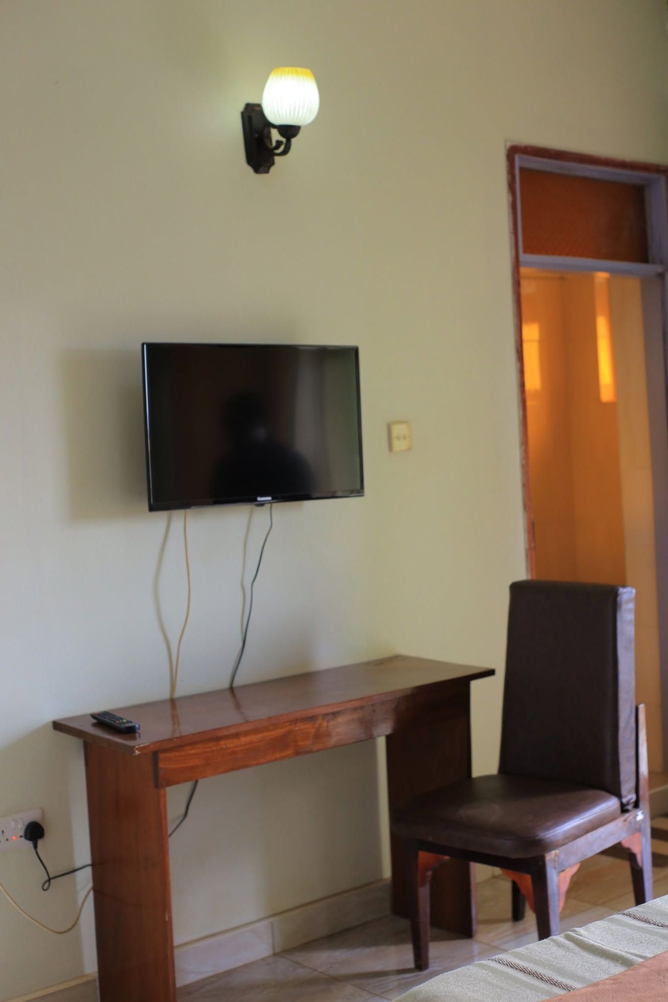 Quarto com televisão de tela plana na parede, escrivaninha de madeira com cadeira e luminária fixada na parede.