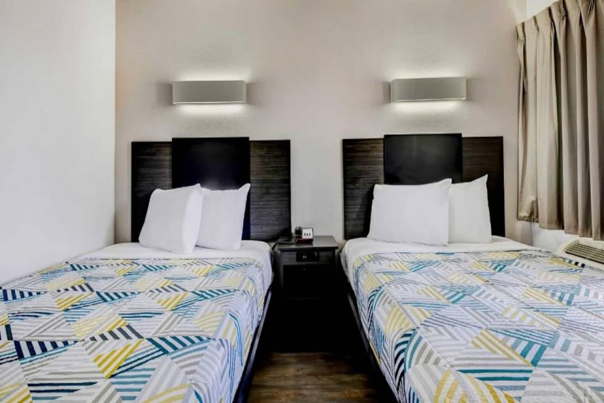 Une chambre d’hôtel avec deux lits, chacun recouvert d’une couvre-lit à motifs, une table de chevet avec une lampe et un luminaire fixé au mur.