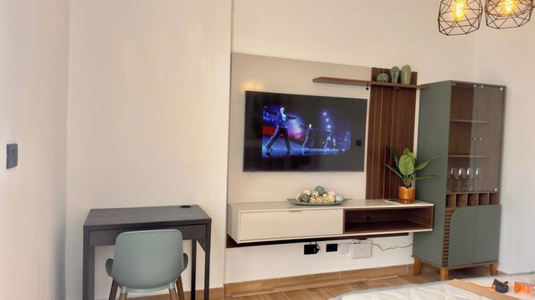 Een moderne woonkamer met een flatscreen televisie aan de muur, een houten kast met glazen deuren, een bureau met een stoel en een potplant.