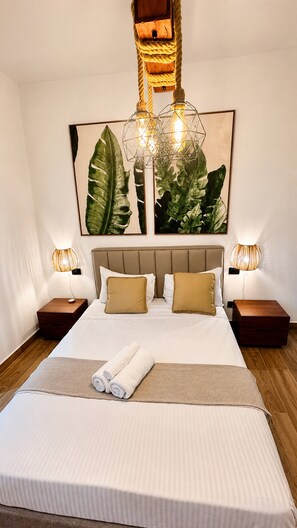 Een netjes opgemaakt bed met hoofdeinde, twee nachtkastjes met lampen, en een grote plant in een lijst aan de muur.
