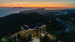A building with a modern design, situated on a hillside overlooking a coastal cityscape at dusk.