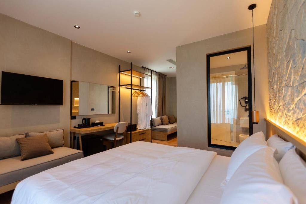 Habitación de hotel con una cama grande, un escritorio, una silla, un sofá, un televisor y un baño visible a través de una puerta de vidrio.