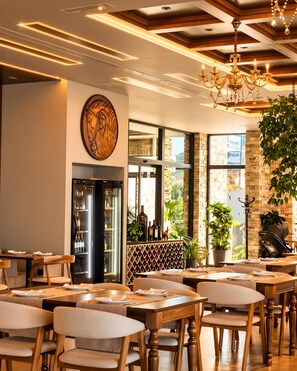 A restaurant interior with wooden tables and chairs, a chandelier, and a wine rack visible through a glass door.