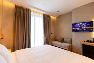 Habitación de hotel con una cama grande, un sofá, un televisor montado en la pared y un ventanal con cortinas.