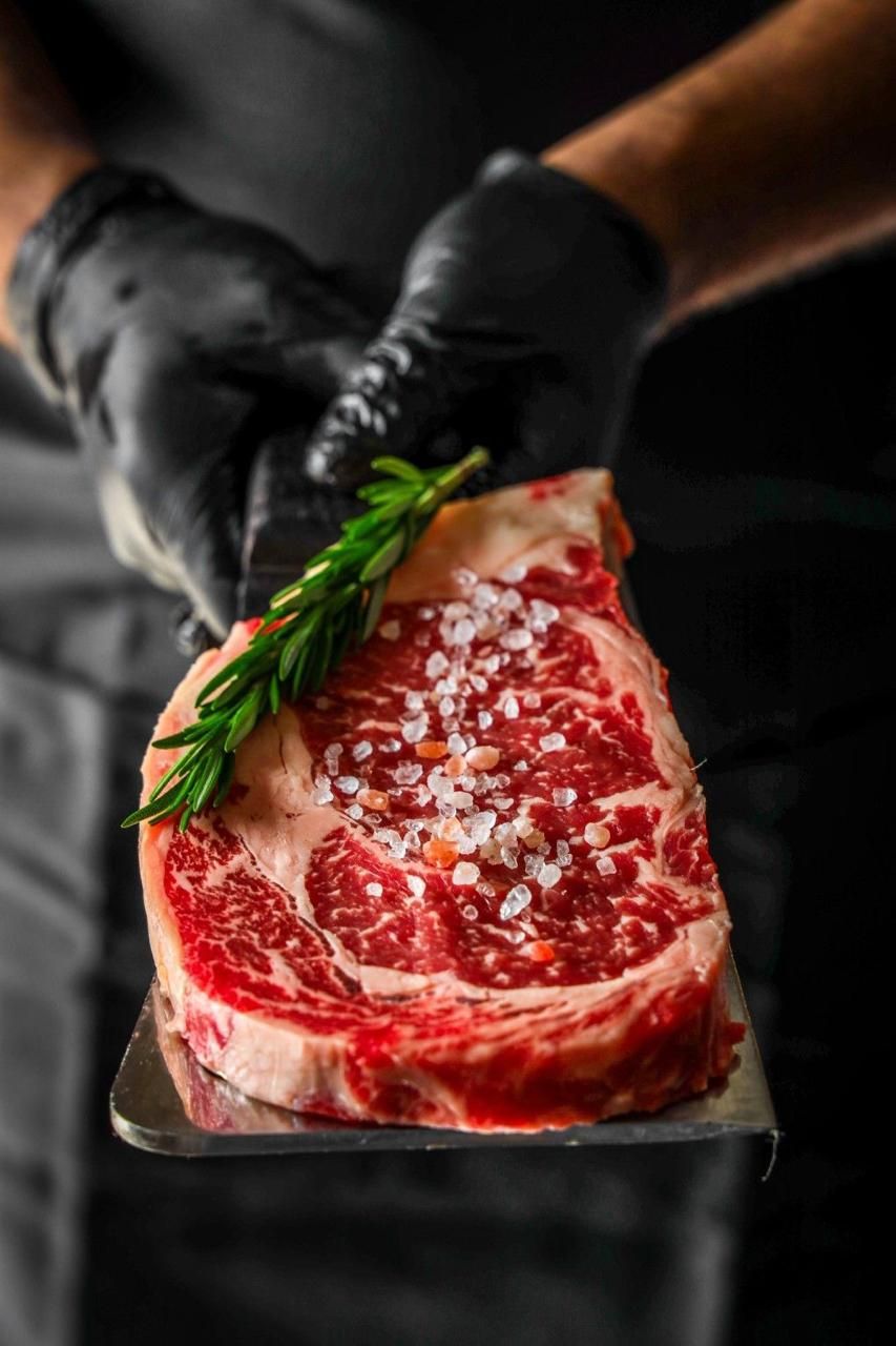 A person holding a steak with a sprig of rosemary and salt on a metal tray.