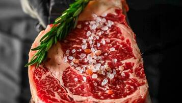A person holding a steak with a sprig of rosemary and salt on a metal tray.