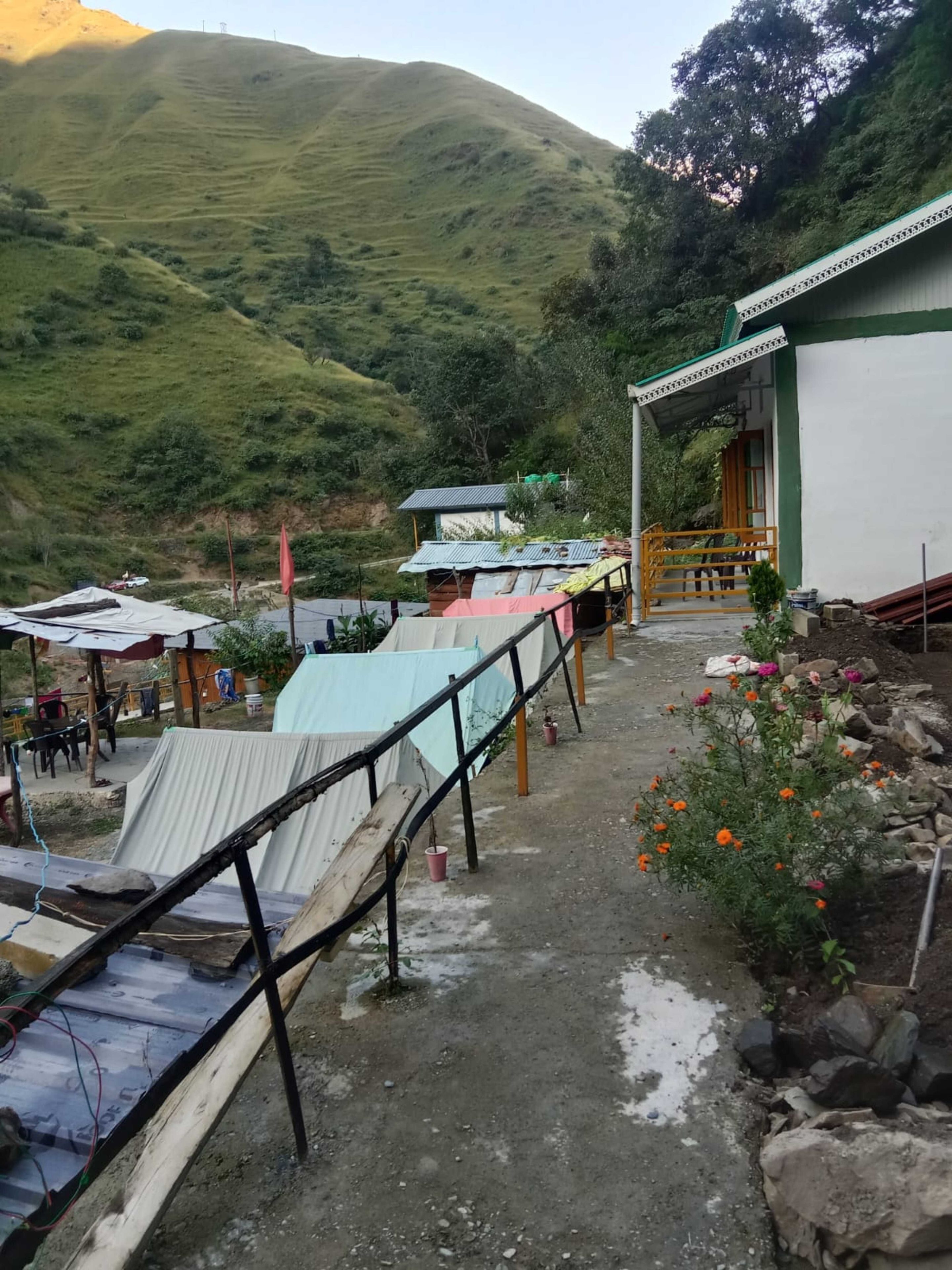A rural setting with a dirt path leading to a building with a green and white exterior. There are makeshift structures and a small garden with flowers.