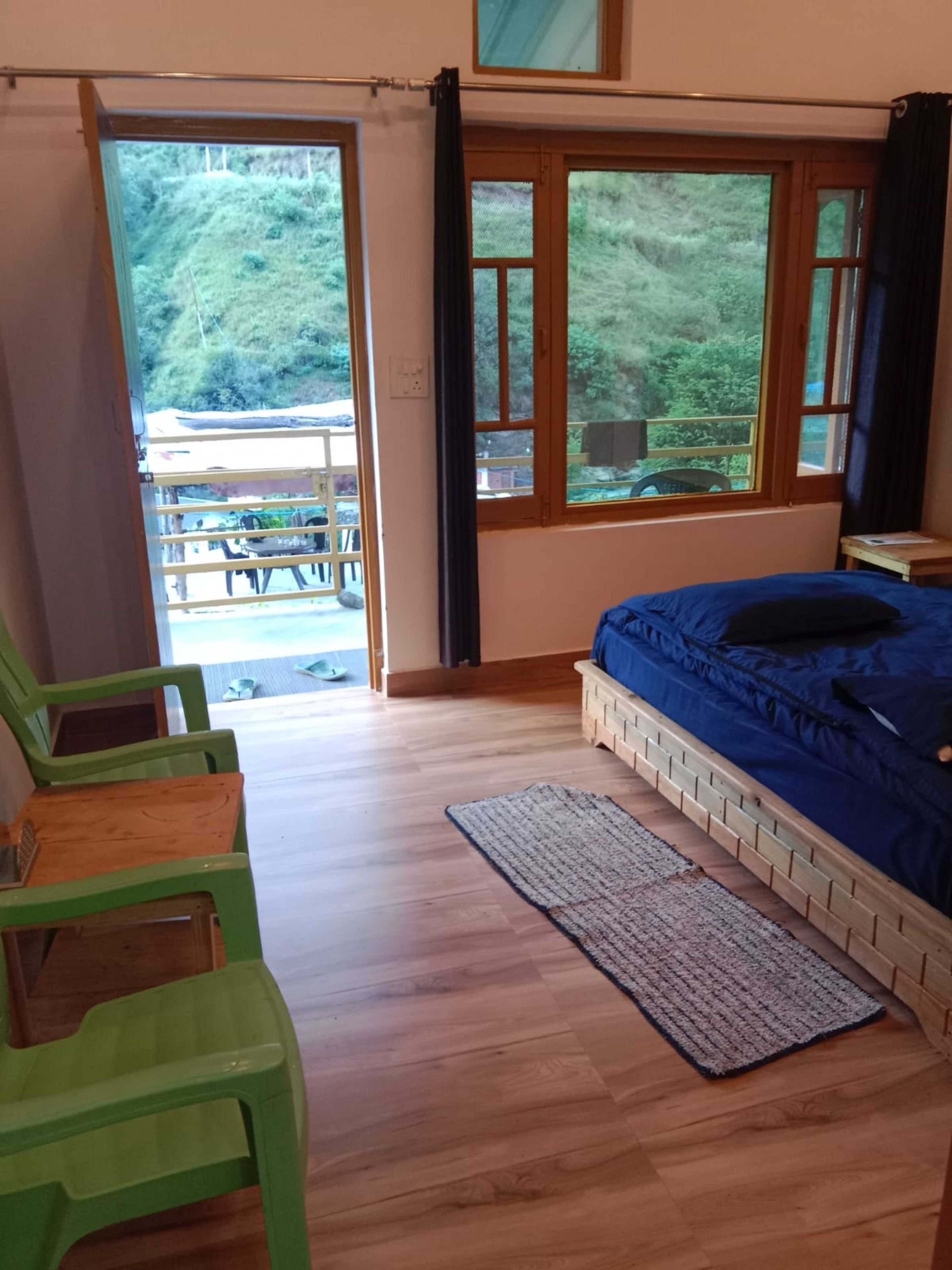 Une chambre avec un lit, un fauteuil vert, une table en bois et une vue sur un balcon aménagé avec du mobilier d’extérieur.