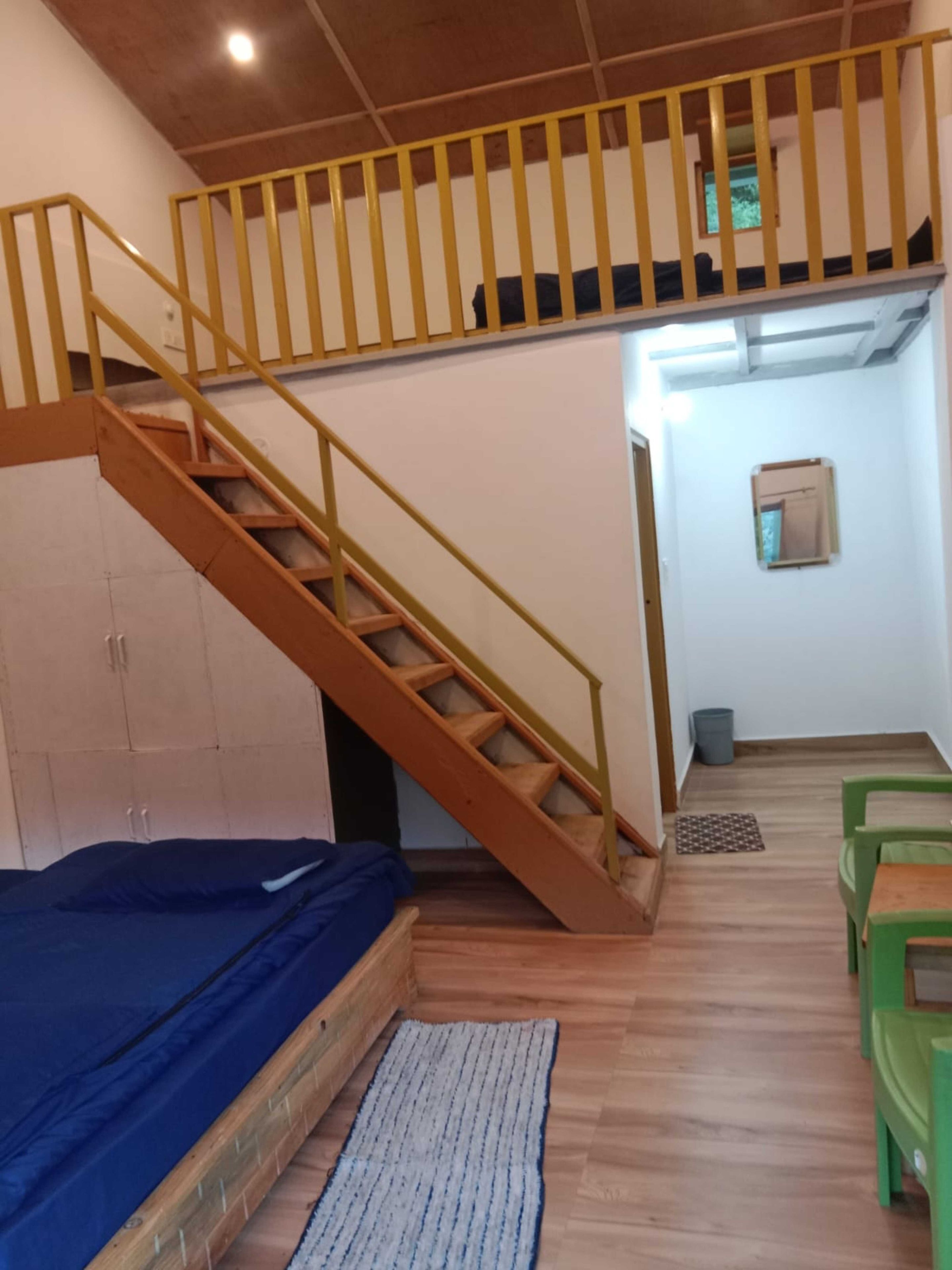 A room with a wooden staircase, a bed, a small table, and green chairs.